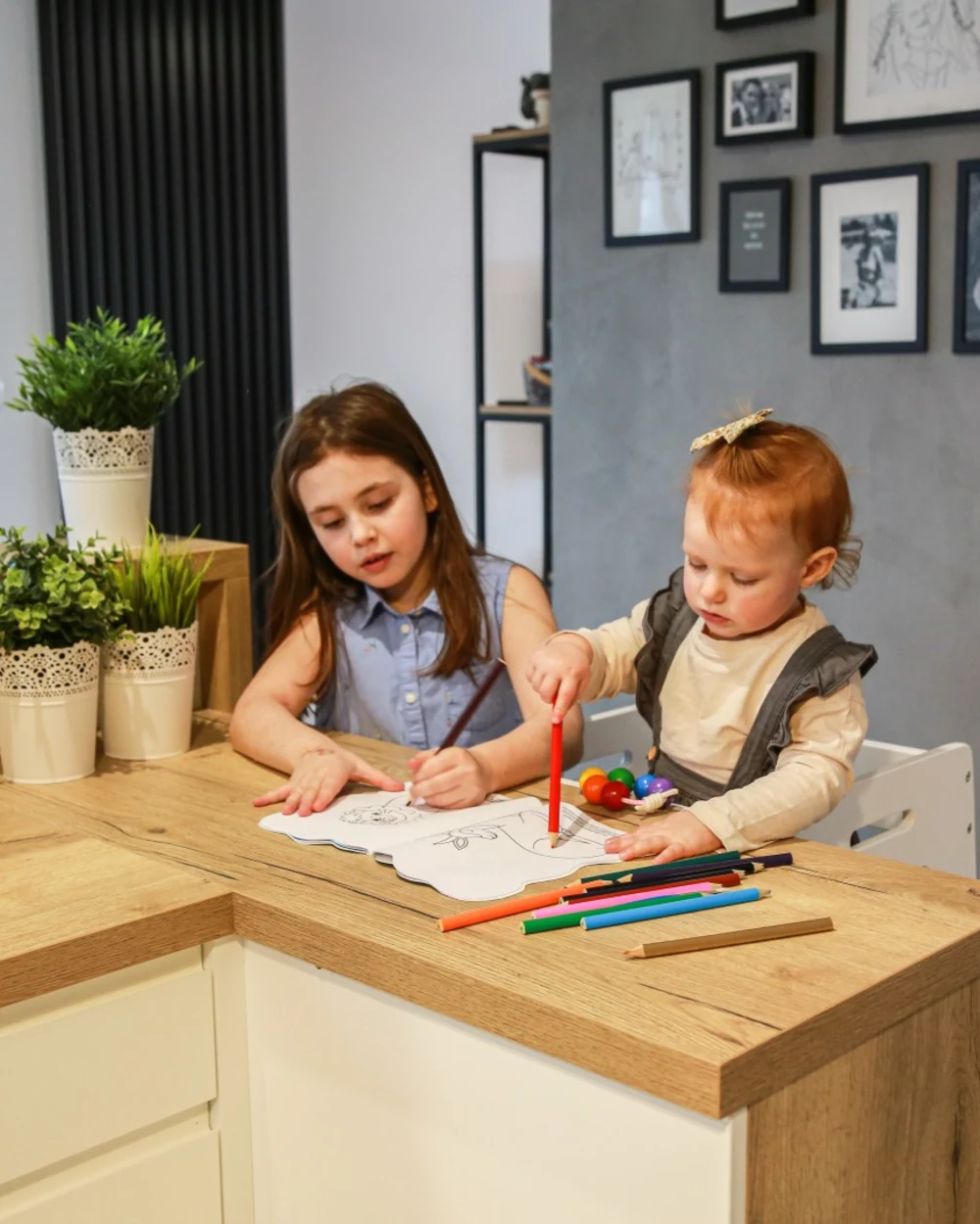 Two siblings drawing at a table, the younger one on a learning tower
